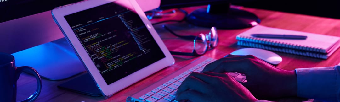 Uma pessoa digitando em um teclado e olhando para um tablet com códigos de programação.